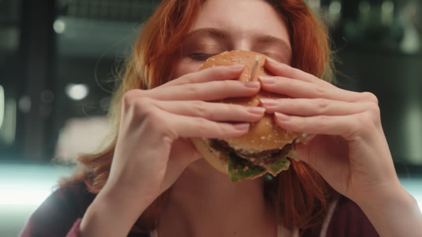 Caucasian woman female girl eating hamburger with eyes closed feeling delight enjoying fast takeaway junk food addiction bad habit meal biting cheeseburger dinner unhealthy snack relaxing at home