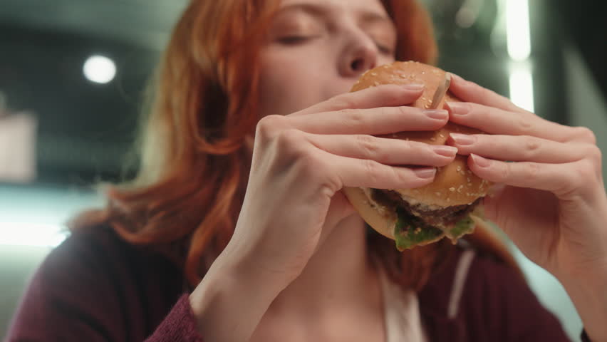 Caucasian woman female girl eating hamburger with eyes closed feeling delight enjoying fast takeaway junk food addiction bad habit meal biting cheeseburger dinner unhealthy snack relaxing at home