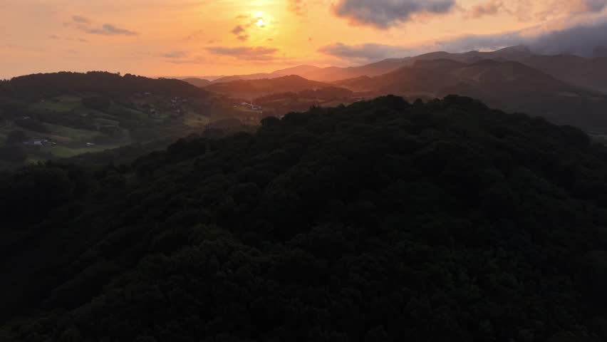 sunset over hilly landscape, glowing sky with dramatic clouds and silhouettes of mountains on horizon.