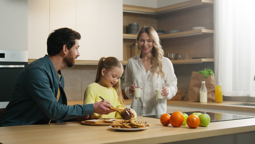 Happy Caucasian family at kitchen at home preparing breakfast together mother hold glasses with milk little daughter child girl prepare toasts bread with chocolate pasta with father help food delivery
