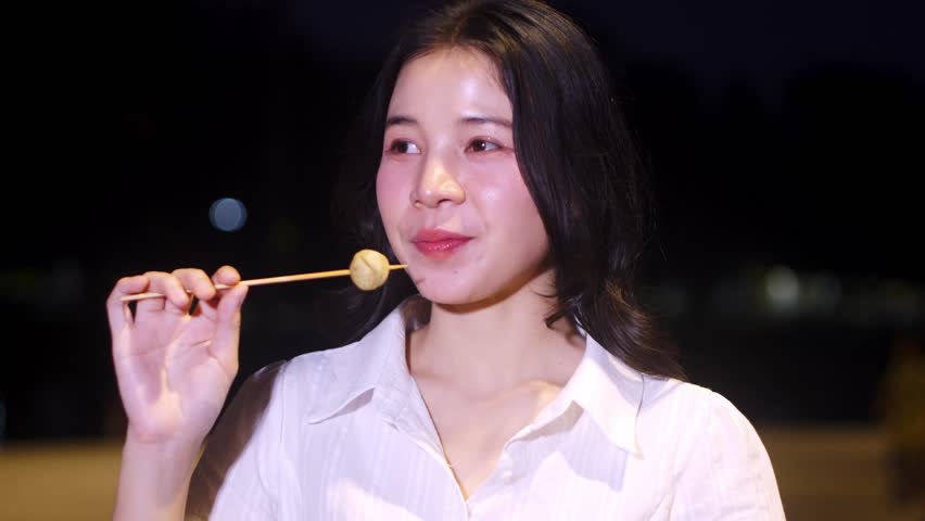 Young woman enjoying street food at night, holding skewered meat on a stick, with city lights softly glowing in the background creating a vibrant atmosphere