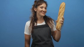 Woman smiling with gloved hands holding a baguette and pointing finger to it in blue studio; cheerful fresh bread. - Powered by Shutterstock - Get 15% off with code: PIKWIZARD15