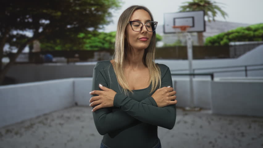 Woman with crossed arms pointing fingers on street near outdoor basketball court wearing glasses and long blonde hair; confidence choice determination.