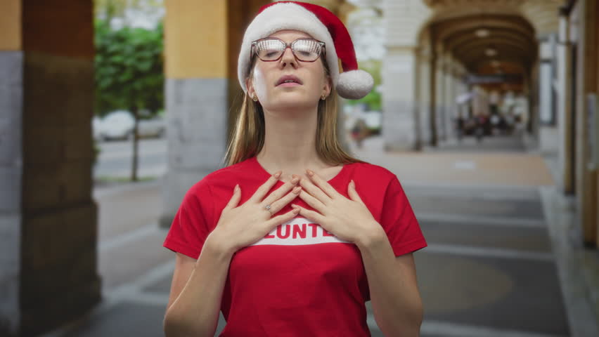 Young woman clutch chest with hands wearing red volunteer shirt and santa hat in outdoor street archway; thankfulness.