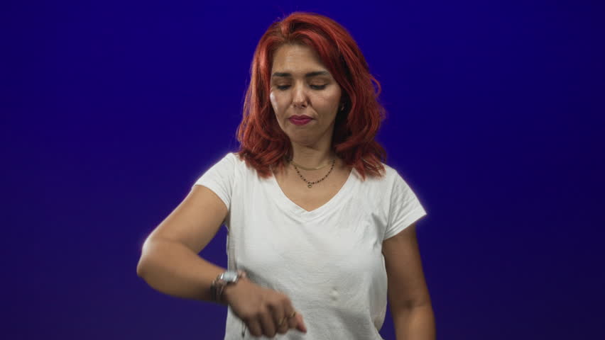 Young woman checking watch and pointing to wrist in studio with deep blue backdrop; impatience time awareness.