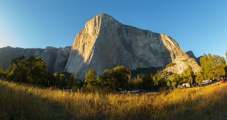 A serene timelapse of sunset over El Capitan in Yosemite National Park, California. The sky transitions from bright blue to warm golden tones, illuminating the iconic granite cliffs in perfect clarity