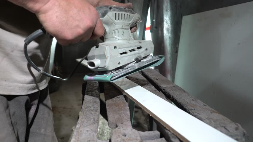 A person is sanding a wooden plank using an electric sander. The workshop is filled with tools and materials, showcasing a crafting activity focused on creating smooth surfaces.