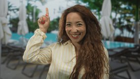 Woman smiling and pointing finger at hotel poolside lounge chairs and umbrellas wearing yellow striped shirt with long curly hair; vacation joy. - Powered by Shutterstock - Get 15% off with code: PIKWIZARD15
