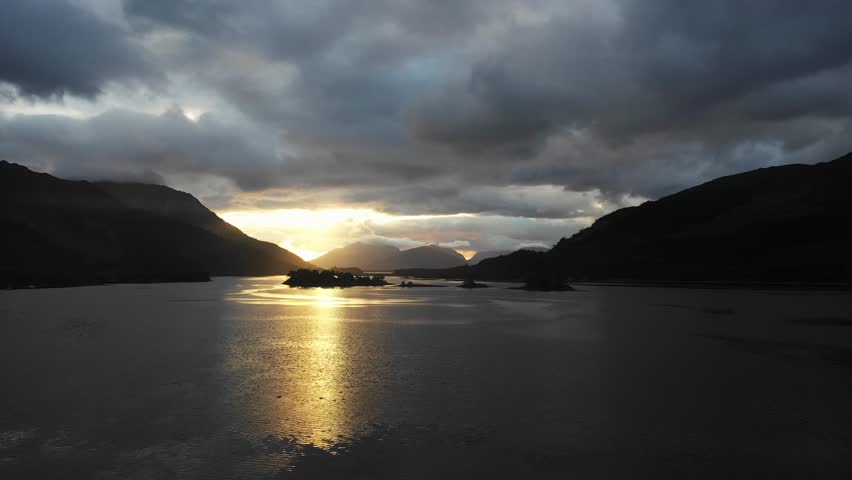 Aerial drone view of Loch Leven in Scotland at sunset, with dramatic clouds over the mountains and sun reflecction on the water - Moody weather in Scottish highlands