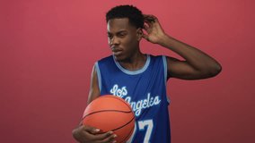 Young man in blue basketball jersey firmly holds basketball and touches ear in red studio; contemplation. - Powered by Shutterstock - Get 15% off with code: PIKWIZARD15