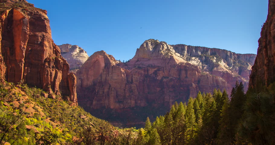 A peaceful daytime timelapse panning across the cliffs of Zion National Park, Utah. Tree shadows shift gently along the red rock walls beneath a clear blue sky, showcasing the calm desert landscape.