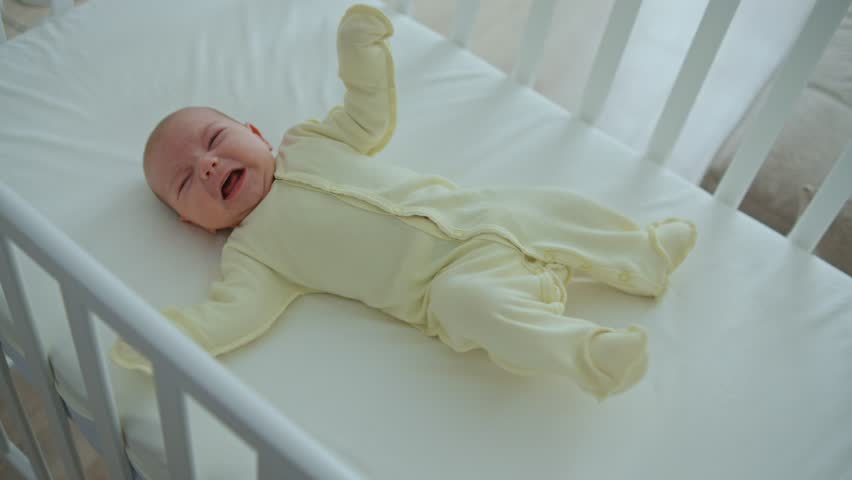 A one-month-old baby lies in a wooden crib, kicking and crying