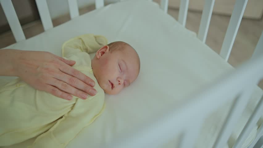 A mother gently strokes a one-month-old baby who is sleeping in a white wooden crib