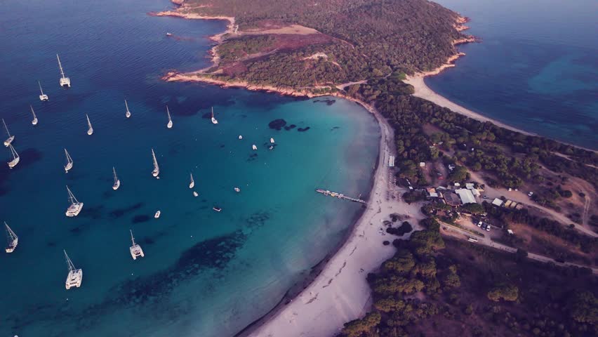 Sailing ships are anchored in a bay on the Mediterranean island of Corsica. A picturesque jetty on the long sandy beach completes the picture. Evening atmosphere.
