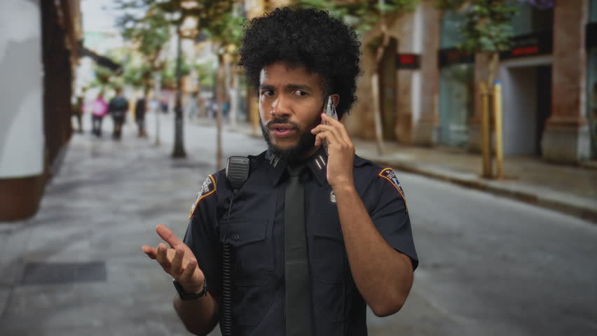 African american police officer in uniform talking on smartphone in a city street setting, illustrating urban communication and law enforcement presence outdoors.