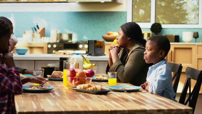 Black children playing around and fighting with food while mom ignores them, woman being disconnected and in burnout due to motherhood and goofy kids. Family serving breakfast at home. Camera B.
