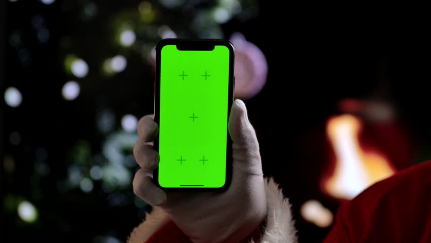 Santa Claus hand in a white glove holds a smartphone with a green screen chroma key in front of a fireplace and a Christmas tree. A warm festive atmosphere for digital or themed mockups. Slow motion.