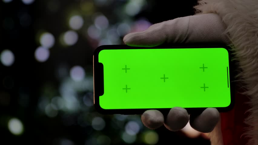 Santa Claus hand in a white glove holds a smartphone with a green screen chroma key in front of a fireplace and a Christmas tree. A warm festive atmosphere for digital or themed mockups. Slow motion.