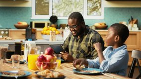 African american dad on tablet multitasking and chatting with his son, serving breakfast with his family and eating good homemade food in the kitchen. Togetherness and joy. Camera B. - Powered by Shutterstock - Get 15% off with code: PIKWIZARD15
