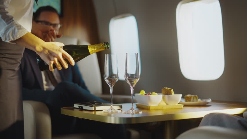 Flight attendant provides luxury service pouring sparkling wine for a successful businessman traveling first class, enjoying a comfortable and exclusive journey on a private airplane
