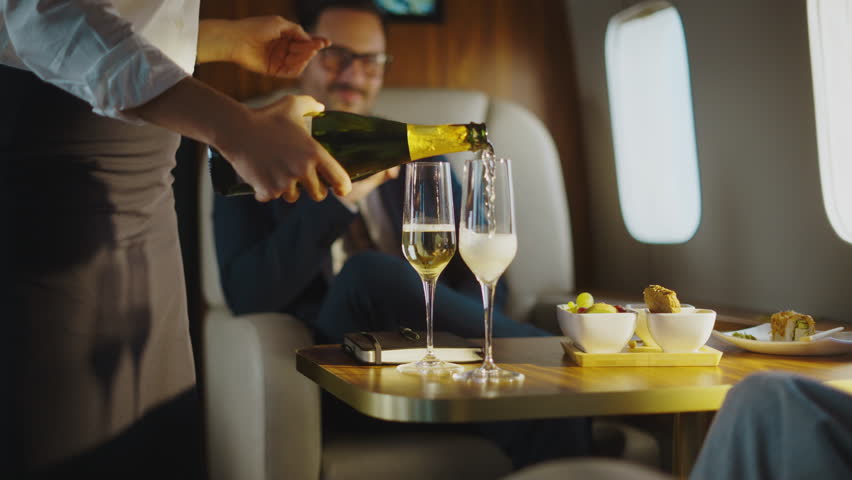 Professional flight attendant pouring sparkling wine into glasses for a successful businessman relaxing in his comfortable seat during a luxurious first-class flight with elite service
