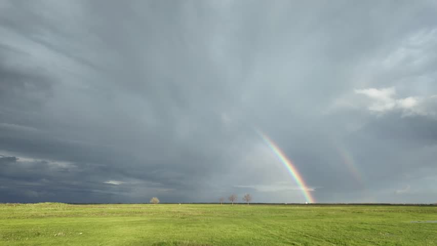 Time-lapse of double rainbow over green meadow under gray storm clouds after rain, cinematic natural landscape