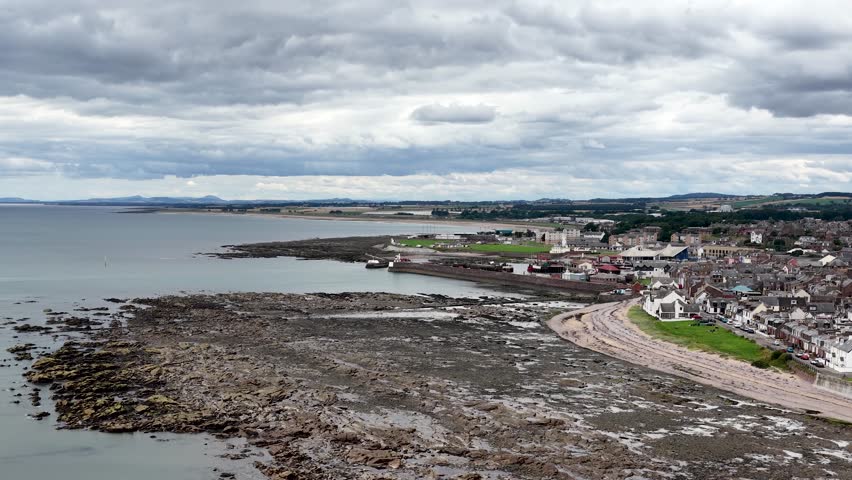 Aerial camera pans across rocky coastline, harbor, and town under cloudy daylight in Dundee, Scotland