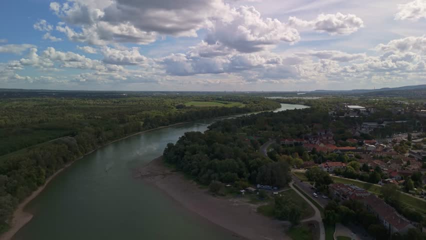 Drone flying forward south along the Danube River at Szentendre outskirts, Hungary, revealing lush forests, riverside curves, and the distant Budapest skyline under dramatic afternoon clouds.