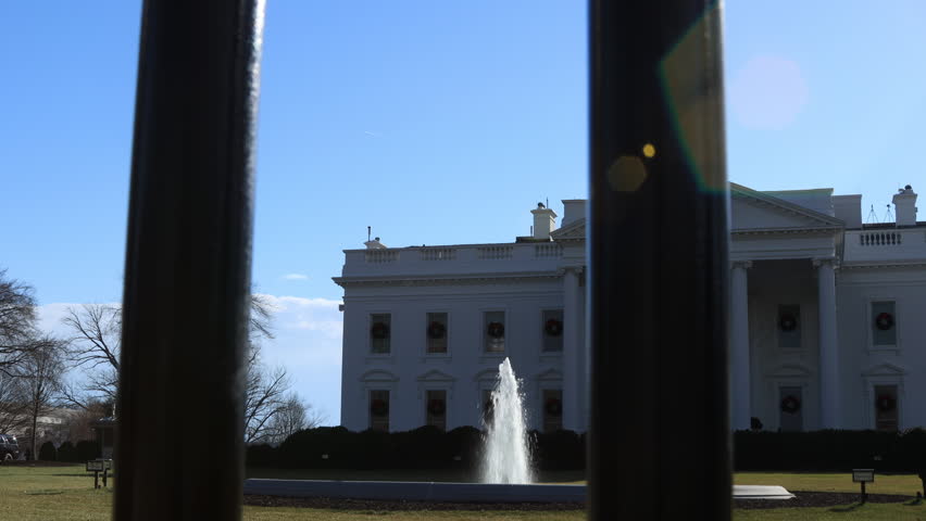 The White House, decorated with Christmas wreaths, is seen through the North Lawn fence. The shot captures the fountain and bright sun flare on a clear day as the camera pans left to right.