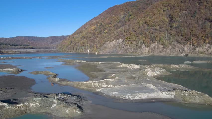 Drone footage of a tailings pond with turquoise water, clay banks, and forested hills covered in bright autumn foliage under a clear blue sky.