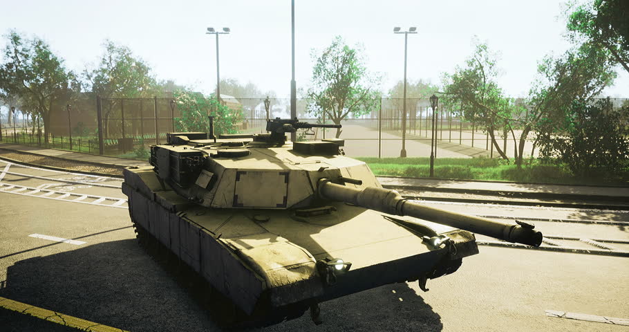 A military tank sits on a paved area adjacent to a fenced sports field. The bright sunlight illuminates the surroundings, creating a vibrant atmosphere.