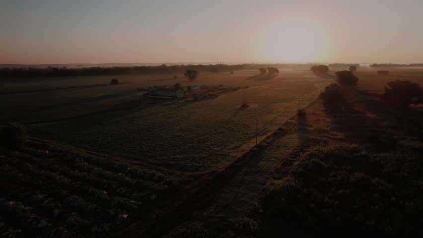 Aerial sunrise over Nebraska farmland near Maxwell, Nebraska. Morning shadows across crop fields and a lone farmhouse. Quiet Great Plains agriculture and rural American livelihood at first light.