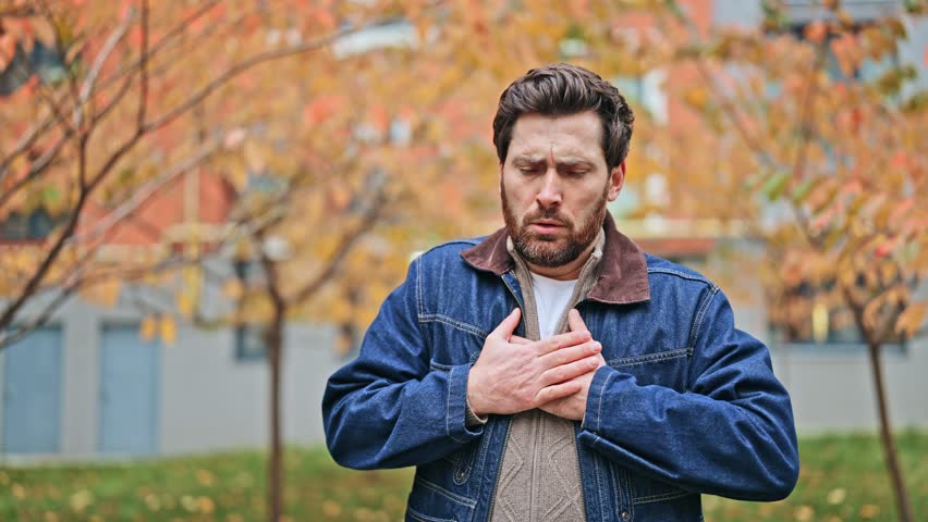 Adult individual stands outdoors experiencing sudden, severe chest pain. Discomfort visible on face, hands pressed over heart. Expresses fear distress and urgency, signaling need for medical attention