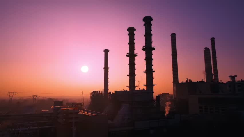 Drone video of industrial factory in Italy silhouetted against vibrant sunset. Smokestacks emit faint plumes under colorful sky, creating a striking, dramatic scene.