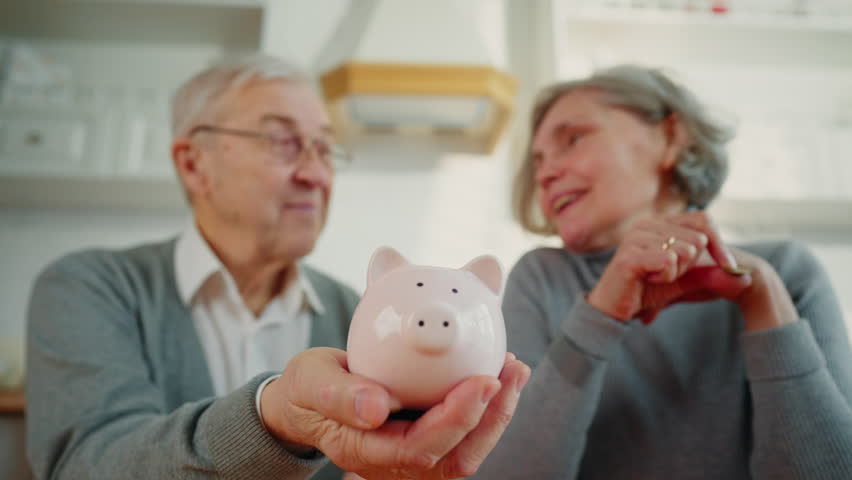 Closeup of piggy bank in hand of aged man when his wife putting coins inside. Elderly people caring about finance and family budget, planning incomes and expenses, joyful pensioners saving money