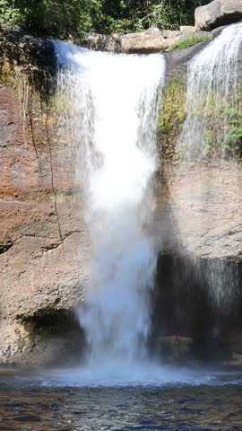Waterfall plunges over rocky cliff into clear pool, surrounded by lush forest in daylight.