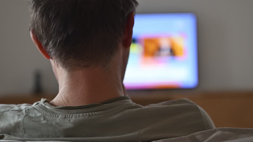 Man sitting on sofa, looking exhausted and uninterested while television glows in background, symbol of boredom and daily monotony. Rear view, 4K with selective focus.