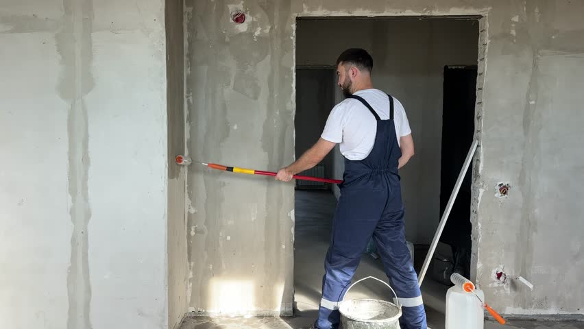 A bearded man in a work uniform is priming the walls after applying gypsum plaster