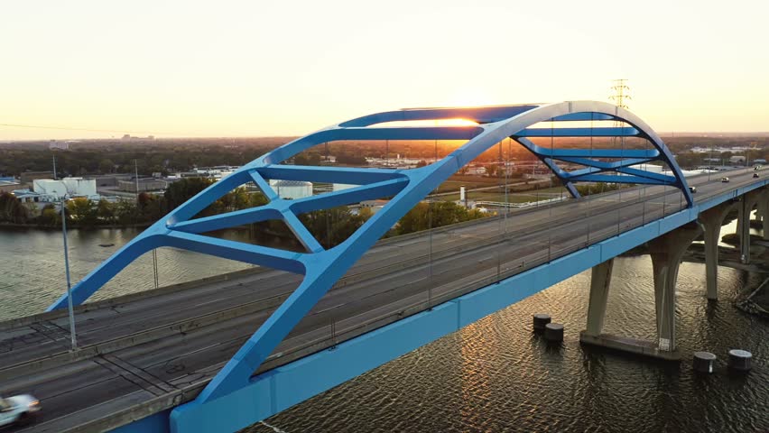 The Leo Frigo Memorial Bridge glows in the warm hues of sunset as cars cross its blue steel frame over the Fox River, reflecting golden light across the calm rippling water.