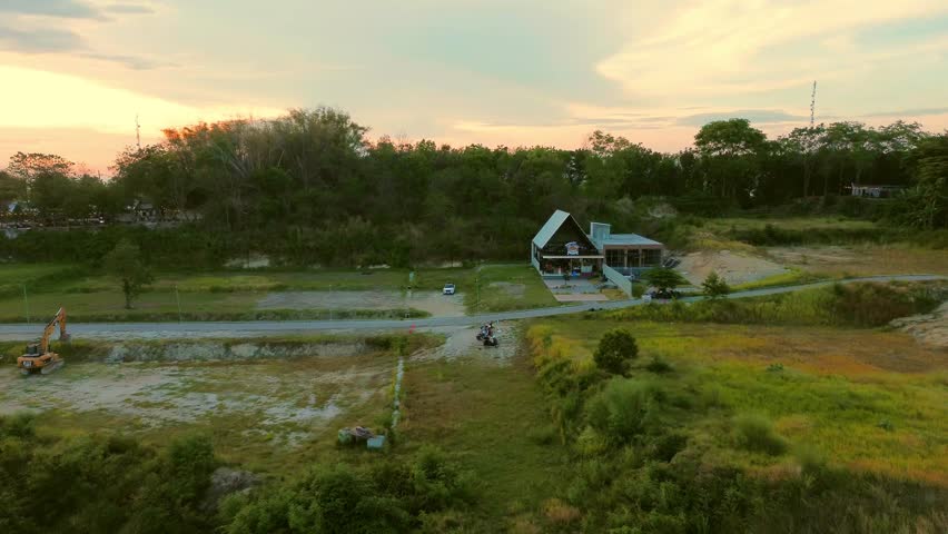 Aerial drone view of a hillside area at sunset, capturing a peaceful landscape with residential houses, lush greenery, and a warm orange sky. Tranquil evening scenery over a city surrounded by hills