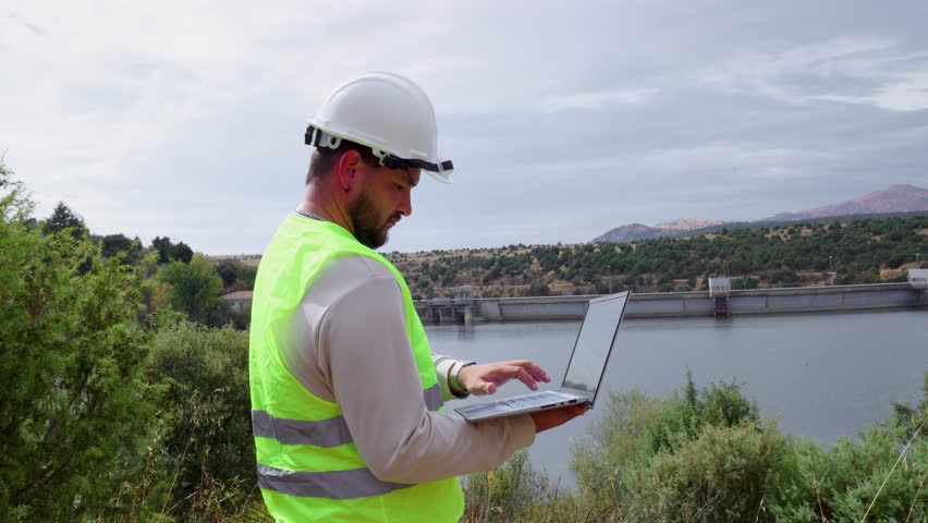 Male engineer in safety gear, including a hard hat and reflective vest, is using a laptop while overseeing the operations of a hydroelectric dam to ensure efficient renewable energy production