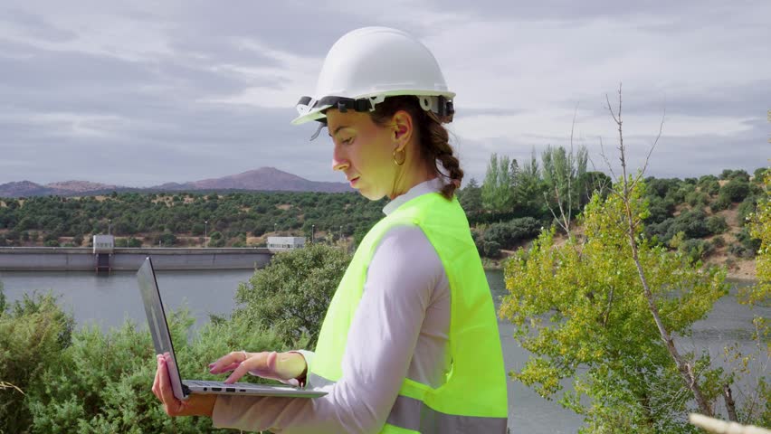 Determined woman engineer wearing a hard hat is utilizing a laptop for maintenance inspection at an outdoor reservoir of a hydroelectric power plant - Powered by Shutterstock - Get 15% off with code: PIKWIZARD15