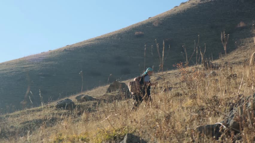 A wide angle lens captures from the side a determined traveler climbing a steep mountain slope with a large backpack and a headband. Traveling in adulthood. High mountain air is beneficial.
