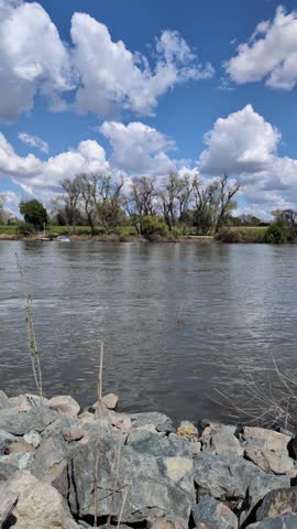A calm river flows beneath bright blue skies dotted with white clouds and distant trees on shore