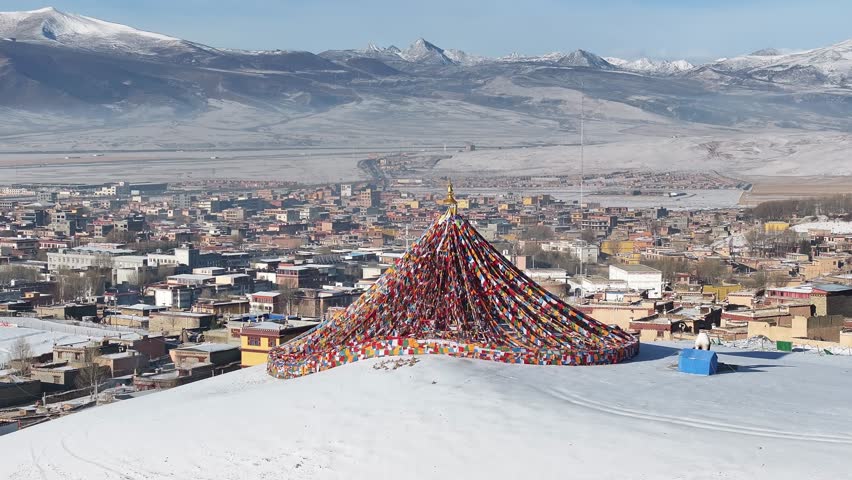 Aerial view of prayer flags cascading down a snowy hill with the city of Litang and snow capped mountains in the background, Litang, China.
