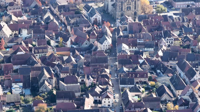 Aerial view of tightly packed buildings and a church tower, the red rooftops contrasting with the autumn trees, Rouffach, Grand Est, France.