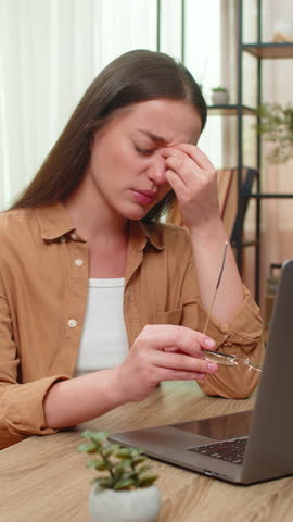 Young woman sitting at home table removes glasses rubs tired eyes after a heavy long laptop work session. Caucasian girl feels physical strain blinking rapidly and seeking rest to ease discomfort now.