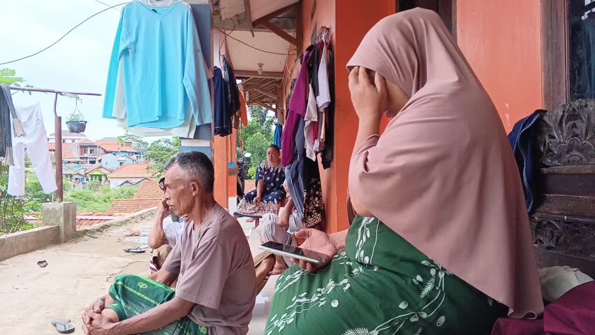 Group of multi-generational Southeast Asian people (men and women) relaxing and socializing on a porch in a crowded rural village setting. Authentic scene of daily community life.