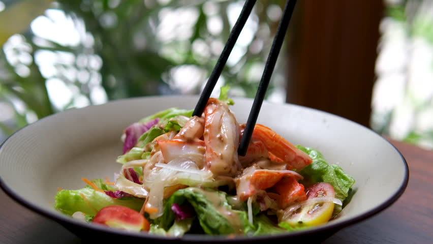 Colorful Chinese cold salad tossed with spicy chili sauce, peanuts, sesame, and chili flakes in a white plate with blue rim, stir-fried with chopsticks for even flavor.