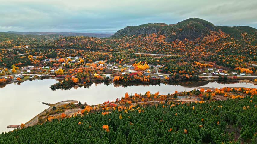 Drone aerial sweeps over the Newfoundland town of North West Brook, showing houses, roads and a lake shimmering with autumn colours beneath a rugged mountain.